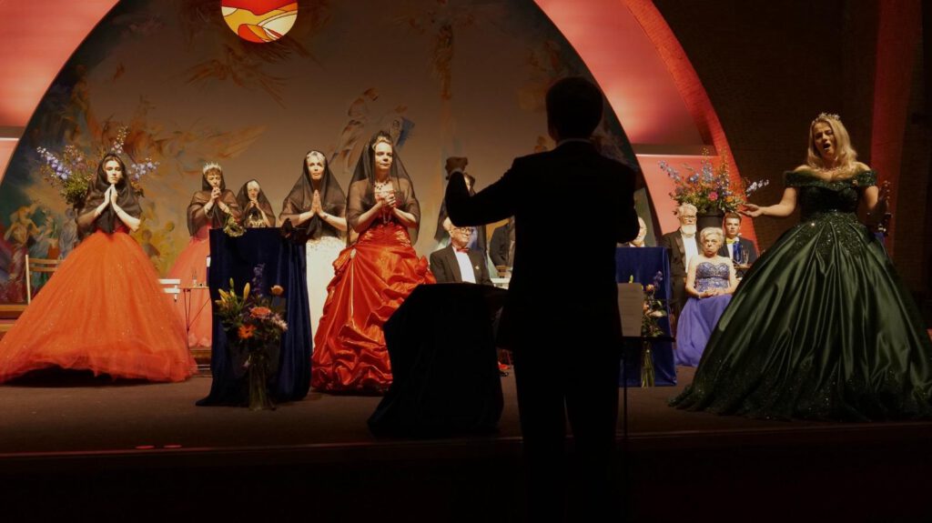 Koor Wilhelmina zingt onder leiding van de dirigent, in een scène met zwart geklede solisten en kleurrijke decors.
