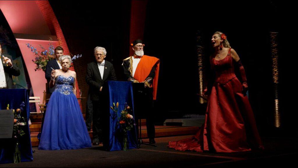 Opname van het openingsconcert met vrouwelijke soliste in rode toga centraal op het podium, begeleid door koorleden.