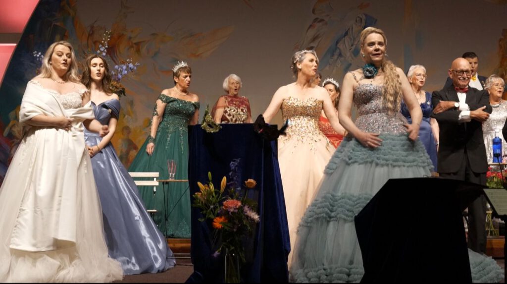 Koor Wilhelmina zingt met passie terwijl vrouwelijke solisten in kleurrijke avondjurken hun partijen uitvoeren.