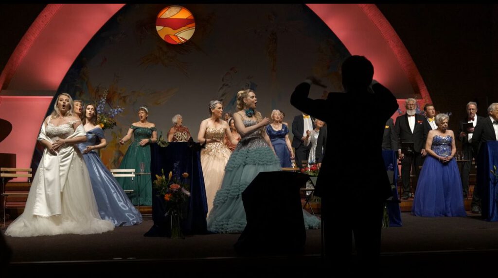 Koor zingt onder begeleiding van dirigent, met de solisten in expressieve houding en kleurrijke aankleding.