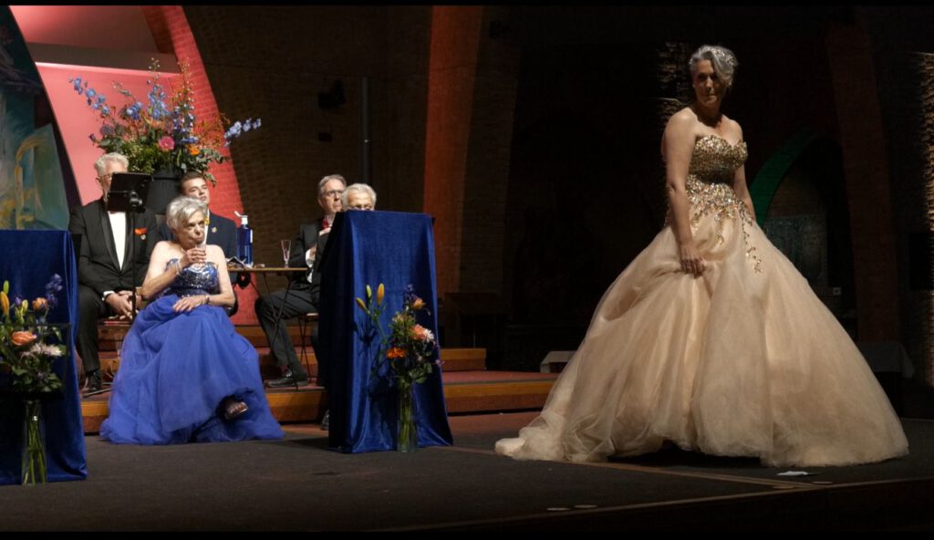 Zangeres in champagnekleurige baljurk staat in stilte op het podium terwijl koorleden aandachtig toekijken.