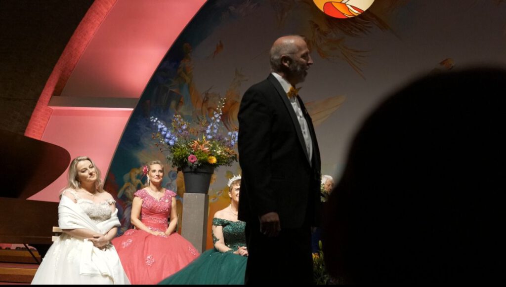 Mannelijke solist loopt over het podium, met op de achtergrond kleurrijk verlichte koorleden in galajaponnen.