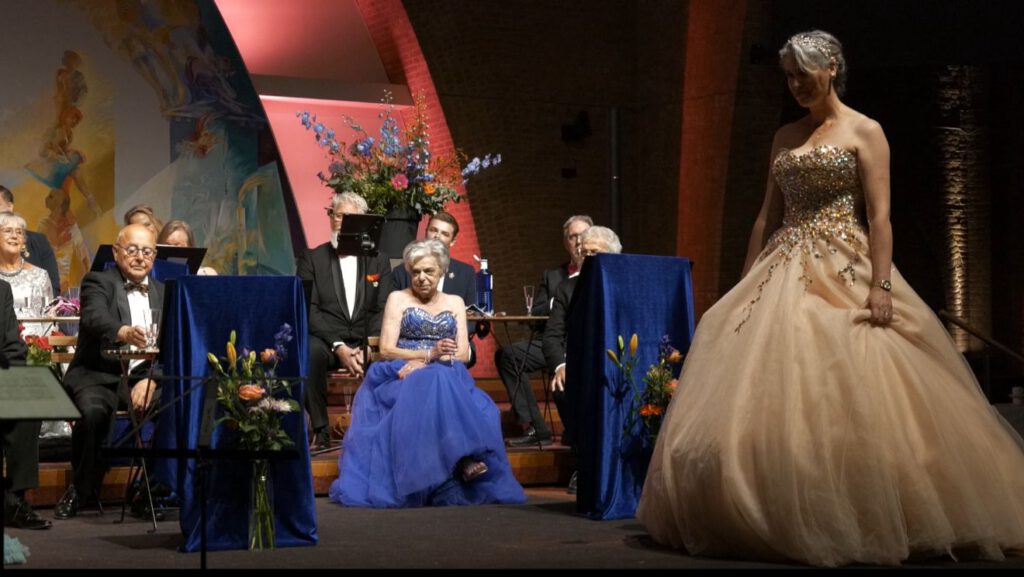 Zangeres in champagnekleurige baljurk staat in stilte op het podium terwijl koorleden aandachtig toekijken.